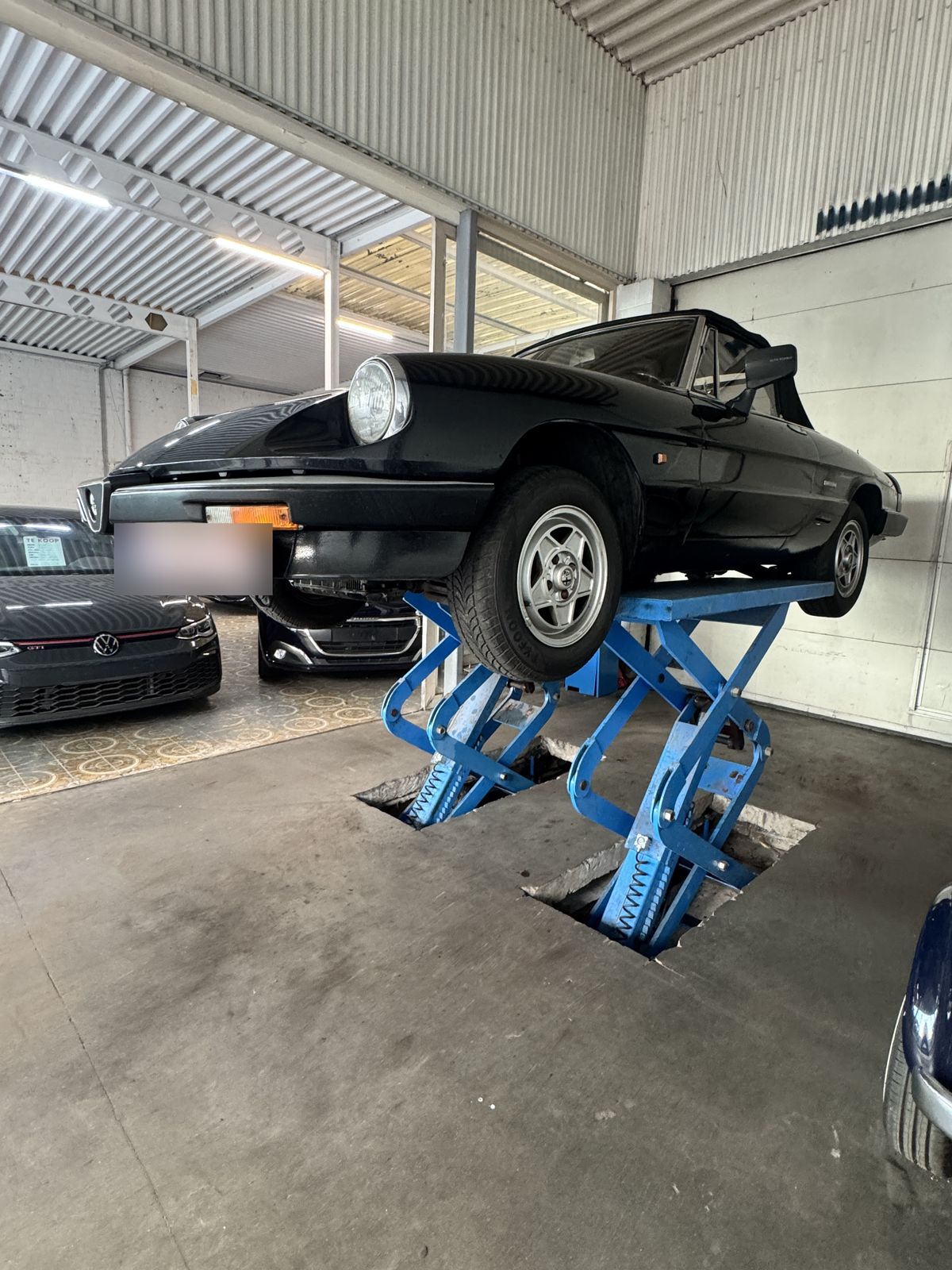 Zwarte Alfa Romeo Spider op een blauwe schaarbrug in het nette werkhuis van JW Motors in Deurne